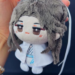Pu&ograve; includere: Una bambola di peluche con corpo bianco, mantello bianco e acconciatura grigia e soffice. La bambola ha occhi marroni, bocca piccola arancione e dettagli rossi. Un nastro bianco &egrave; attaccato alla bambola.