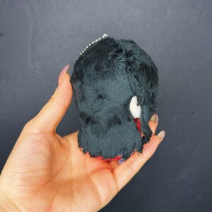 Pu&ograve; includere: Una piccola creatura nera di peluche con dettagli bianchi e rossi. Il giocattolo fatto a mano ha una consistenza soffice e una piccola catena d'argento attaccata in cima. La creatura &egrave; tenuta in una mano su uno sfondo scuro.