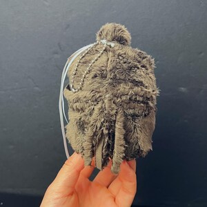 Pu&ograve; includere: Un ornamento fatto a mano, marrone e peloso, con un piccolo pompon in cima. L'ornamento ha nastri bianchi e una catena di perline d'argento. La parte inferiore dell'ornamento ha diverse strisce di tessuto pendenti.