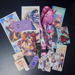 May include: A collection of Genshin Impact merchandise, including character art prints, a lanyard, stickers, a keychain, and a business card. The items feature vibrant colors and various character designs from the popular video game.