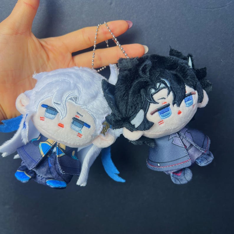 May include: Two plush dolls with anime-style features. One doll has white hair and blue accents, the other has black hair and a dark outfit. Both dolls have blue eyes and are attached to a silver chain.