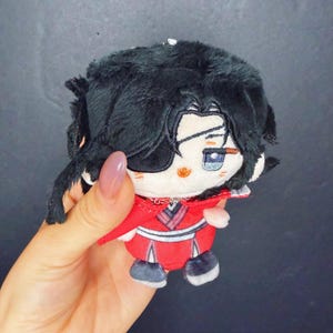 Pu&ograve; includere: Una bambola di peluche con capelli neri e una benda sull'occhio. La bambola indossa un completo rosso con dettagli bianchi e scarpe nere. La bambola &egrave; tenuta in una mano.