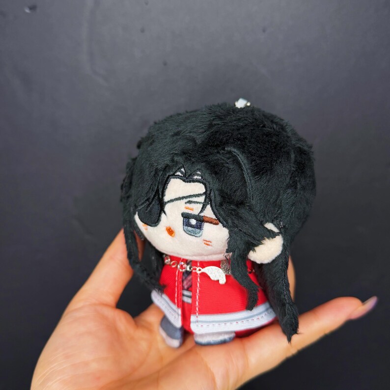 Pu&ograve; includere: Una bambola di peluche con la testa rotonda e capelli neri e soffici. La bambola indossa un abito rosso con dettagli bianchi e una catena d'argento. Il viso della bambola ha occhi blu e un'espressione neutra. La bambola &egrave; tenuta in una mano.