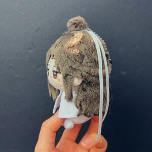 Pu&ograve; includere: Una bambola di peluche con capelli di pelliccia marrone e un vestito bianco. La bambola ha una testa piccola e rotonda con tratti del viso ricamati, tra cui occhi marroni e una bocca piccola. Nastri bianchi pendono dalla parte superiore della bambola.