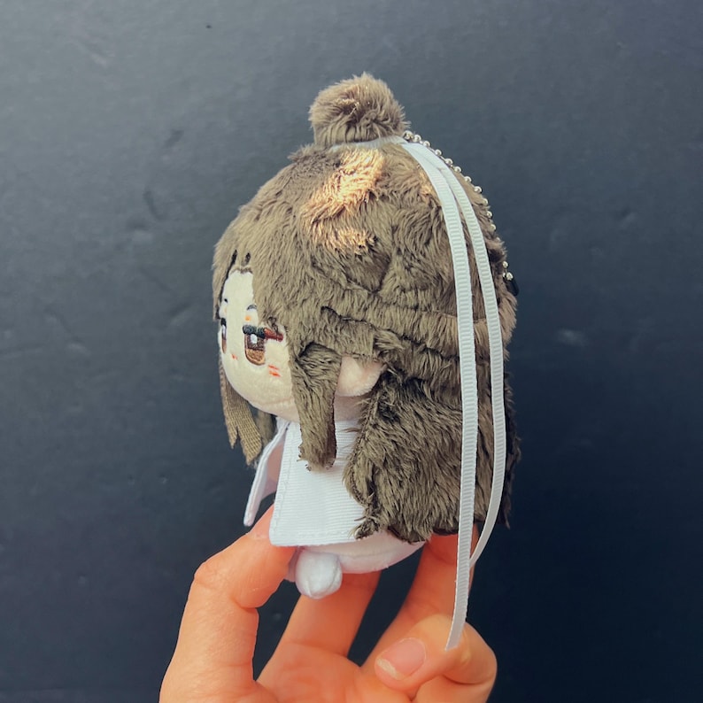 Pu&ograve; includere: Una bambola di peluche con capelli di pelliccia marrone e un vestito bianco. La bambola ha una testa piccola e rotonda con tratti del viso ricamati, tra cui occhi marroni e una bocca piccola. Nastri bianchi pendono dalla parte superiore della bambola.