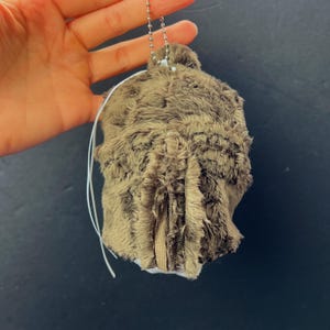 Pu&ograve; includere: Un portachiavi in ​​peluche marrone con una catena d'argento e un filo bianco. Il peluche &egrave; un materiale morbido e strutturato con una cerniera sul fondo. Il portachiavi &egrave; tenuto su uno sfondo scuro.