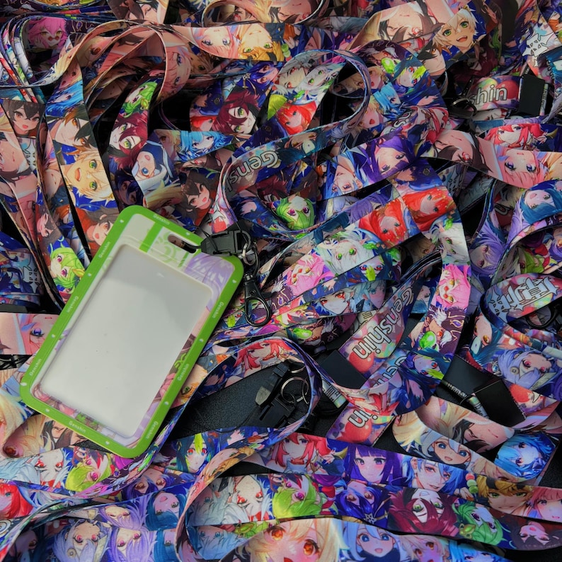 May include: A pile of colorful lanyards featuring anime-style character illustrations. The lanyards are predominantly blue, purple, and pink, with a variety of character designs. A green and white ID holder with a clear window is also present.