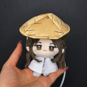 Pu&ograve; includere: Una bambola di peluche con capelli castani, che indossa un cappotto bianco e un cappello color cachi. La bambola ha tratti del viso ricamati, tra cui occhi marroni e una piccola bocca arancione. La bambola &egrave; tenuta in mano su sfondo nero.