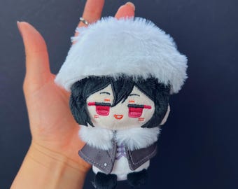 Può includere: Una bambola di peluche con capelli neri, dettagli rossi e un cappello e colletto di pelliccia bianchi. La bambola indossa una giacca grigia sopra una camicia bianca con un motivo a quadri. La bambola è di piccole dimensioni.