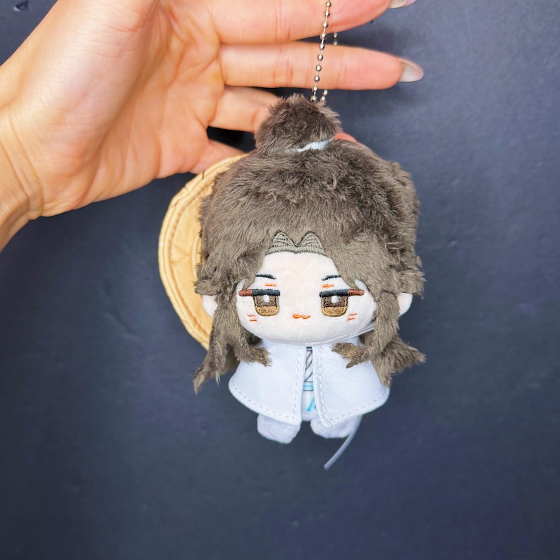 Pu&ograve; includere: Una bambola di peluche con capelli castani e un cappotto bianco, attaccata a un portachiavi. La bambola ha un viso dettagliato con occhi marroni e tiene in mano un piccolo oggetto rotondo di colore marrone chiaro. I capelli della bambola sono acconciati a chignon.