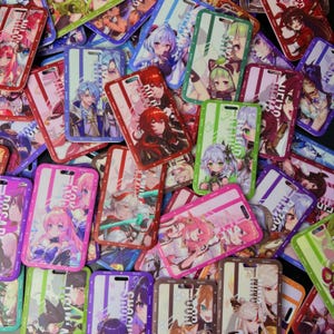May include: A collection of rectangular phone cases with colorful anime character designs. The cases feature various characters in vibrant colors, with a mix of pink, green, blue, and red accents. Each case has a rectangular shape with a cutout for a lanyard.