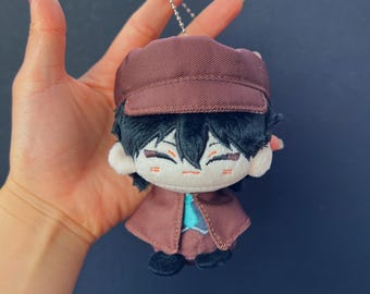 Può includere: Bambola di peluche di un personaggio con cappello e cappotto marrone, con capelli neri e un viso sorridente. La bambola ha una piccola catena d'argento attaccata in alto. L'outfit della bambola include una camicia blu.
