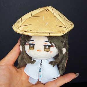 Pu&ograve; includere: Una bambola di peluche con un cappello di paglia e lunghi capelli castani. La bambola ha un cappotto bianco e un viso dettagliato con occhi marroni e fard. La bambola &egrave; tenuta in mano su uno sfondo scuro.
