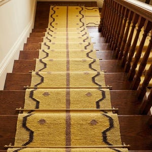 May include: A stair runner with a yellow background and a dark brown border and center stripe. The runner has a repeating pattern and is installed on wooden stairs with a wooden handrail.