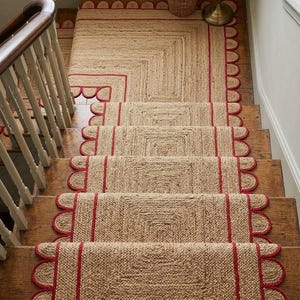 May include: Jute staircase runner rugs in natural tan with a red scalloped edge. The rugs are laid on wooden stairs, providing a coordinated and stylish appearance for the home. The stairs are made of wood.