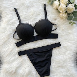 Puede incluir: Un conjunto de lencería negro con un sujetador con ribete de volantes y tirantes ajustables, un bandeau a juego y un tanga. El conjunto se exhibe sobre una superficie blanca y esponjosa, con rosas blancas en el fondo.