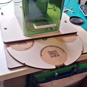 Puede incluir: Un recipiente verde transparente con un asa plateada descansa sobre una base de madera clara. Debajo, se ven dos posavasos semicirculares con centros de corcho y texto impreso y códigos QR. El texto dice: www.kiwidreamsdesigns.nz.