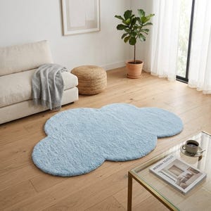 May include: A pale blue, cloud-shaped rug made of soft, fluffy material. The rug is on a wooden floor in a living room setting, with a sofa, ottoman, and coffee table visible. The rug adds a whimsical touch to the room's decor.