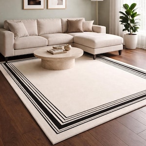 May include: A cream-colored rectangular rug with a black border is the focal point of a living room. The rug is placed on a dark brown hardwood floor. A beige sectional sofa, a round coffee table, and framed artwork complete the room's decor.