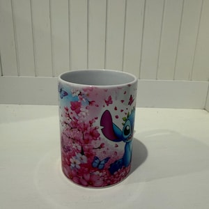 Puede incluir: Una taza de cer&aacute;mica blanca con un estampado gr&aacute;fico colorido de un personaje alien&iacute;gena azul rodeado de vibrantes flores de cerezo rosadas y peque&ntilde;as mariposas azules. El dise&ntilde;o envuelve la superficie exterior del recipiente.