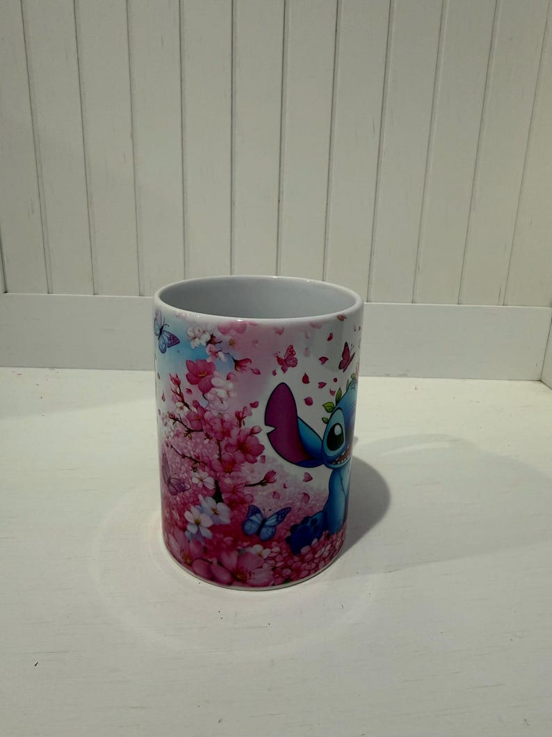 Puede incluir: Una taza de cer&aacute;mica blanca con un estampado gr&aacute;fico colorido de un personaje alien&iacute;gena azul rodeado de vibrantes flores de cerezo rosadas y peque&ntilde;as mariposas azules. El dise&ntilde;o envuelve la superficie exterior del recipiente.