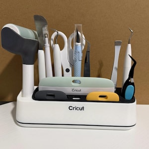 May include: A white Cricut desktop organizer holding various crafting tools including a brayer, scissors, weeding hooks, a craft knife, and tweezers. The organizer features a black interior tray with slots for tools and the Cricut brand name printed on the front.