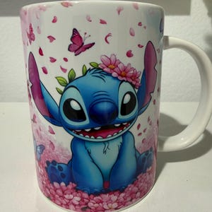 Puede incluir: Una taza de cer&aacute;mica blanca con un gr&aacute;fico de un personaje alien&iacute;gena azul de orejas grandes, que lleva una corona de flores rosas. El personaje est&aacute; rodeado de p&eacute;talos de cerezo rosas, hojas verdes y peque&ntilde;as mariposas sobre fondo blanco.