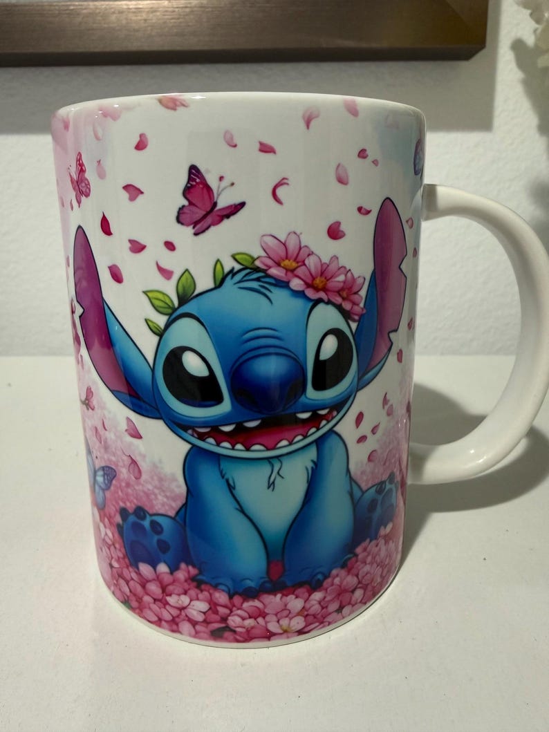 Puede incluir: Una taza de cer&aacute;mica blanca con un gr&aacute;fico de un personaje alien&iacute;gena azul de orejas grandes, que lleva una corona de flores rosas. El personaje est&aacute; rodeado de p&eacute;talos de cerezo rosas, hojas verdes y peque&ntilde;as mariposas sobre fondo blanco.