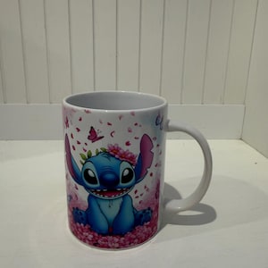 Puede incluir: Una taza de cer&aacute;mica blanca con un personaje alien&iacute;gena azul de orejas grandes y una corona de flores rosas. El dise&ntilde;o incluye un fondo de p&eacute;talos de cerezo rosas cayendo y peque&ntilde;as mariposas, con el personaje sentado entre flores rosas.