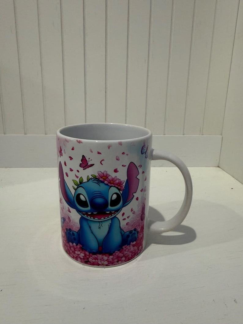 Puede incluir: Una taza de cer&aacute;mica blanca con un personaje alien&iacute;gena azul de orejas grandes y una corona de flores rosas. El dise&ntilde;o incluye un fondo de p&eacute;talos de cerezo rosas cayendo y peque&ntilde;as mariposas, con el personaje sentado entre flores rosas.