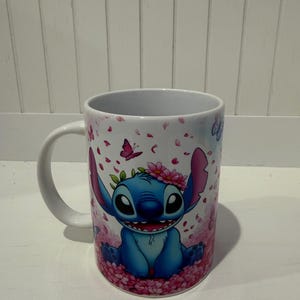 Puede incluir: Una taza de cer&aacute;mica blanca con un personaje alien&iacute;gena azul de orejas grandes y amplia sonrisa. El personaje lleva una corona de flores rosas y est&aacute; rodeado por un patr&oacute;n de p&eacute;talos de cerezo rosas que caen y peque&ntilde;as mariposas moradas.