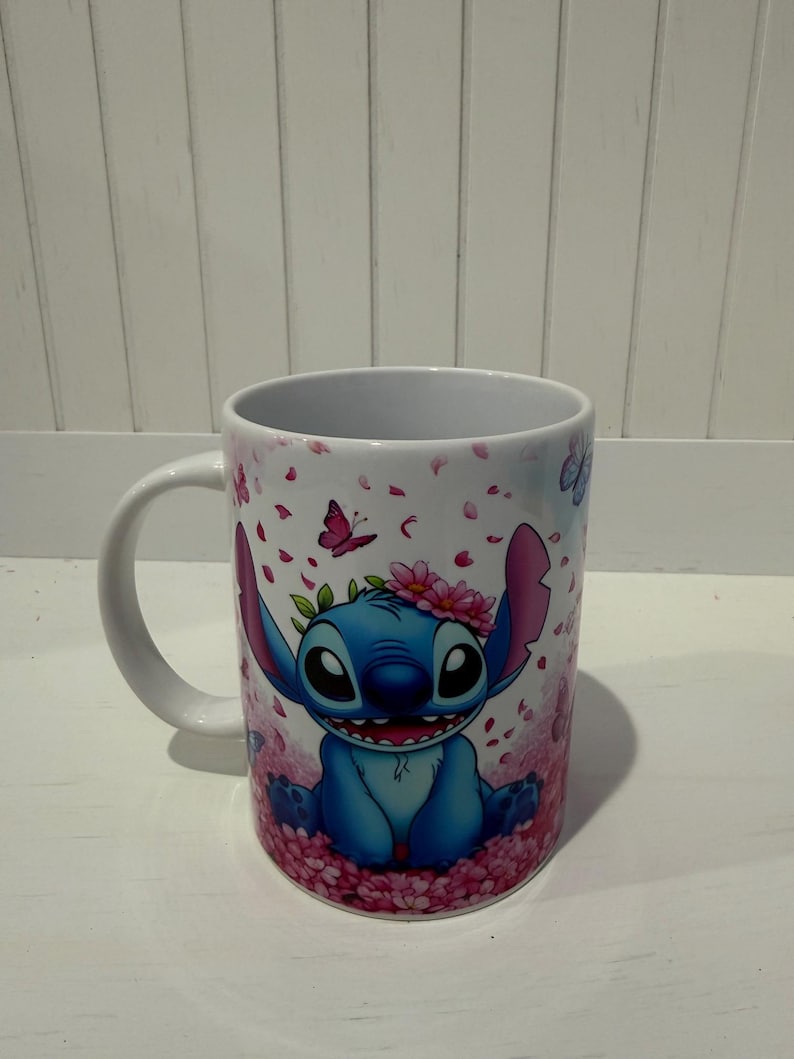 Puede incluir: Una taza de cer&aacute;mica blanca con un personaje alien&iacute;gena azul de orejas grandes y amplia sonrisa. El personaje lleva una corona de flores rosas y est&aacute; rodeado por un patr&oacute;n de p&eacute;talos de cerezo rosas que caen y peque&ntilde;as mariposas moradas.