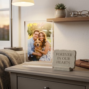 Può includere: Una cornice in acrilico trasparente che mostra una fotografia a colori di una coppia e un golden retriever. Una piccola pietra grigia con la scritta "FOREVER IN OUR HEARTS" è a destra. Su un comodino grigio.