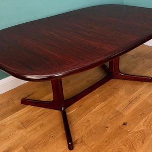 May include: A dark reddish-brown wooden dining table with an oval top and rounded edges. The table features a central support structure with two angled legs connected by a horizontal crossbar, showcasing a smooth, polished wood grain finish.