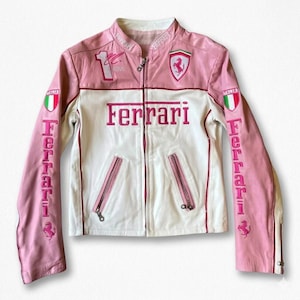 Puede incluir: Chaqueta de cuero Ferrari rosa y blanca con cremallera. Presenta el logotipo de Ferrari, la palabra "Ferrari" en rosa y parches de la bandera italiana. Las mangas tienen la palabra "Ferrari" verticalmente.