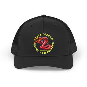 May include: Black trucker cap with a red cobra graphic emitting a laser beam, surrounded by yellow text reading "LASER COBRAS! THEY'RE COBRAWESOME!". The website lasercobras.com is also visible. The back of the cap is mesh.