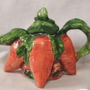 May include: A ceramic teapot shaped like a cluster of orange carrots with green leafy tops. The lid is designed to look like carrot greens, and the handle is formed by a curved green leaf. The surface is glossy, featuring orange and green colors.