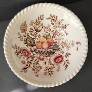 May include: Cream-colored ceramic bowl with a detailed floral and fruit design. The center features a basket overflowing with grapes, peaches, and other fruits, surrounded by flowers and foliage in shades of brown, pink, and green. The rim has a textured, rope-like border.