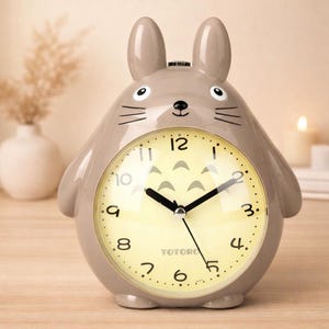 May include: A gray, cartoon-style alarm clock shaped like a character with large ears. The clock face is yellow with black numbers and hands. The word "TOTORO" is printed on the clock face.