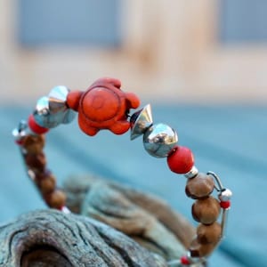 Pode incluir: Uma pulseira de contas com uma conta central laranja em forma de tartaruga, intercalada com contas metálicas prateadas em forma de cone e esfera, contas redondas vermelhas menores e contas de madeira castanha texturizada num fio.