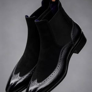 Könnte beinhalten: Ein Paar schwarze Chelsea-Boots aus einer Kombination von glattem, poliertem Leder und weichem Wildleder. Die Stiefel verfügen über dekorative Brogue-Details an der Flügelkappe und eine Zuglasche an der Ferse mit violettem Innenfutter.