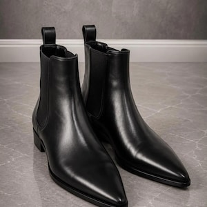 Könnte beinhalten: Ein Paar schwarze Chelsea-Boots aus Leder mit spitzer Zehenpartie, niedrigem Blockabsatz und elastischen Seiteneinsätzen. Jeder Stiefel verfügt über eine vertikale Zuglasche am hinteren Schaftrand und besticht durch eine glatte Oberfläche.