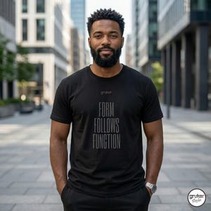 Può includere: T-shirt nero con la scritta "FORM FOLLOWS FUNCTION" in verticale. La maglietta presenta anche il marchio "gruker" in alto. La persona indossa un orologio e si trova di fronte a uno sfondo urbano.
