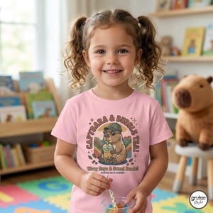 Può includere: Una maglietta rosa con un disegno di capibara che beve bubble tea, con la scritta "CAPYBARA & BUBBLE TEA" e "Wavy Days & Sweet Reads!". La maglietta è indossata da un bambino sorridente.