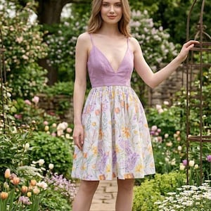 May include: A knee-length dress featuring a solid lavender bodice with a deep V-neckline and thin spaghetti straps. The skirt is white with a colorful floral print in shades of yellow, orange, purple, and green, featuring a gathered waistline.