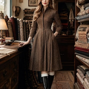 May include: A brown herringbone patterned dress with a high collar, three-quarter length sleeves, and a pleated skirt that falls below the knee. The dress features a front button closure and side pockets, paired with black knee-high leather boots.