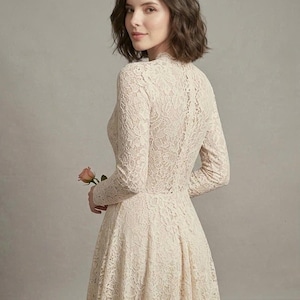 May include: A long-sleeved dress made of cream-colored floral lace featuring a high neckline and a fitted bodice that transitions into a flared skirt. The intricate lace pattern covers the entire garment, which is worn with a single pink rose.