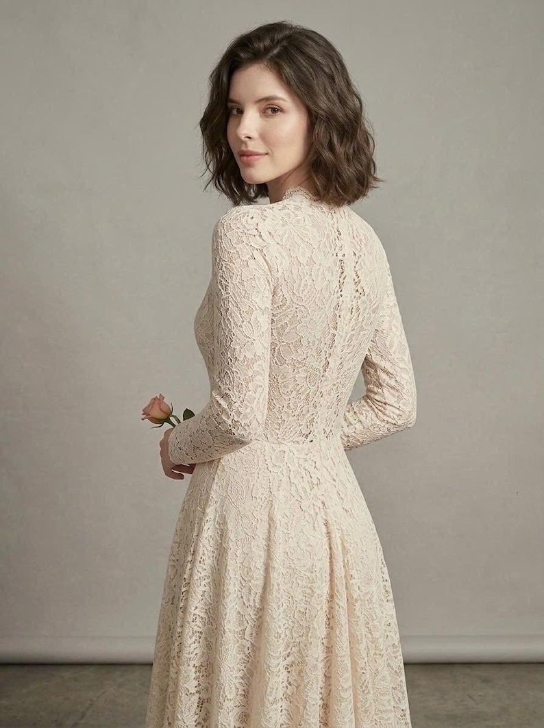 May include: A long-sleeved dress made of cream-colored floral lace featuring a high neckline and a fitted bodice that transitions into a flared skirt. The intricate lace pattern covers the entire garment, which is worn with a single pink rose.