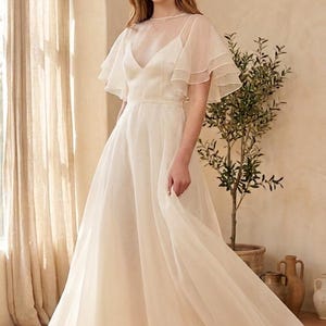 May include: A floor-length cream-colored gown featuring a sheer overlay, a V-neckline, and layered flutter sleeves. The dress has a fitted bodice that transitions into a flowing, full-length skirt made of lightweight, semi-sheer fabric.