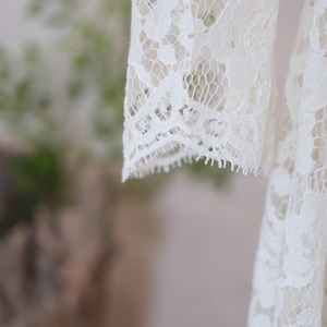 May include: An off-white lace garment featuring long sleeves with intricate floral patterns and scalloped edges. The delicate fabric displays a detailed openwork design throughout, highlighting the craftsmanship of the lace texture and the hem.